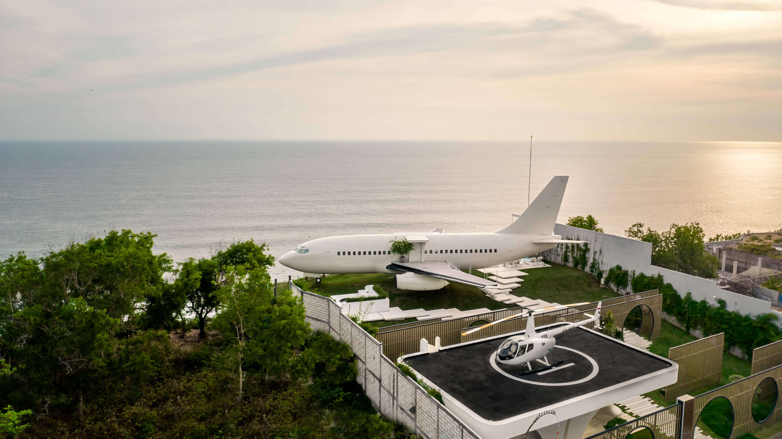The Private Jet Villa helipad on the cliff edge at sunset, with a helicopter on the pad and the Boeing 737 beyond it